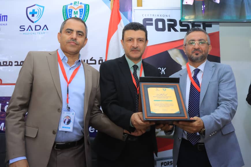 Three men in suits at an award ceremony; the man in the center holds a framed certificate presented by the man on the right, all wearing red lanyards.