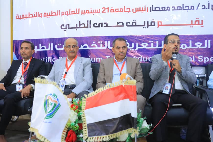 Panel of four men in suits seated on a stage during a conference, with one man speaking into a microphone; Arabic banner backdrop and national flag props on the table in front.