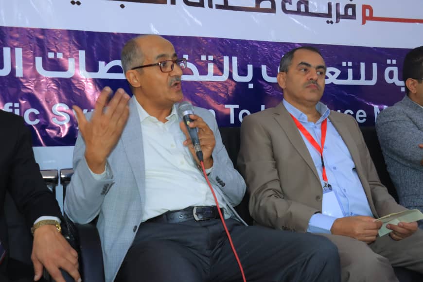 Panel of three men on stage; the man in a light blazer speaks into a microphone while others listen, with a banner in Arabic behind them.