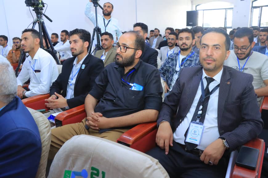 Audience of men seated in red chairs at a conference, wearing lanyards and badges, with camera crew in the background.
