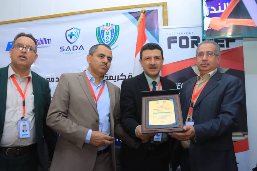 Four men in suits at a ceremony, one holding a framed award plaque, with banners in the background and badges on lanyards front.
