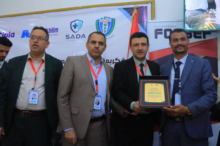 Four men in suits at an awards ceremony, one presenting a framed certificate to a recipient, with banners in the background in a conference setting.