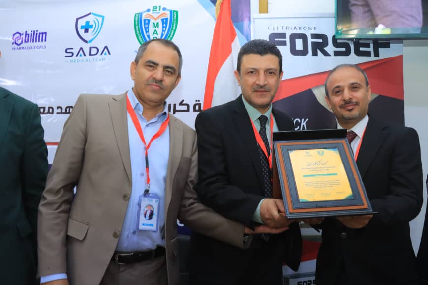 Three men in suits pose on stage as the man in the center presents a framed certificate to the man on the right, with banners and a flag in the background.