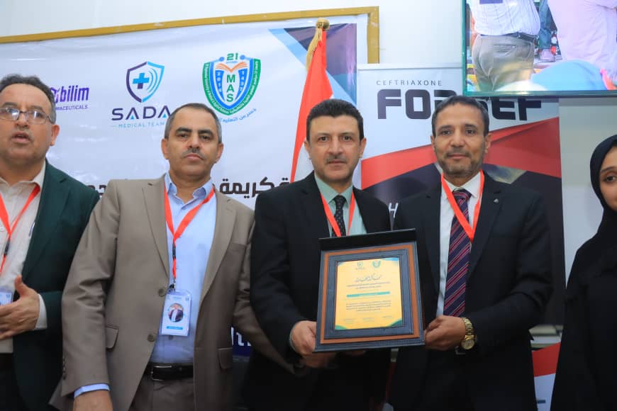 Five professionals in suits at an award ceremony; the man in the center holds a framed certificate, with banners and logos in the background.