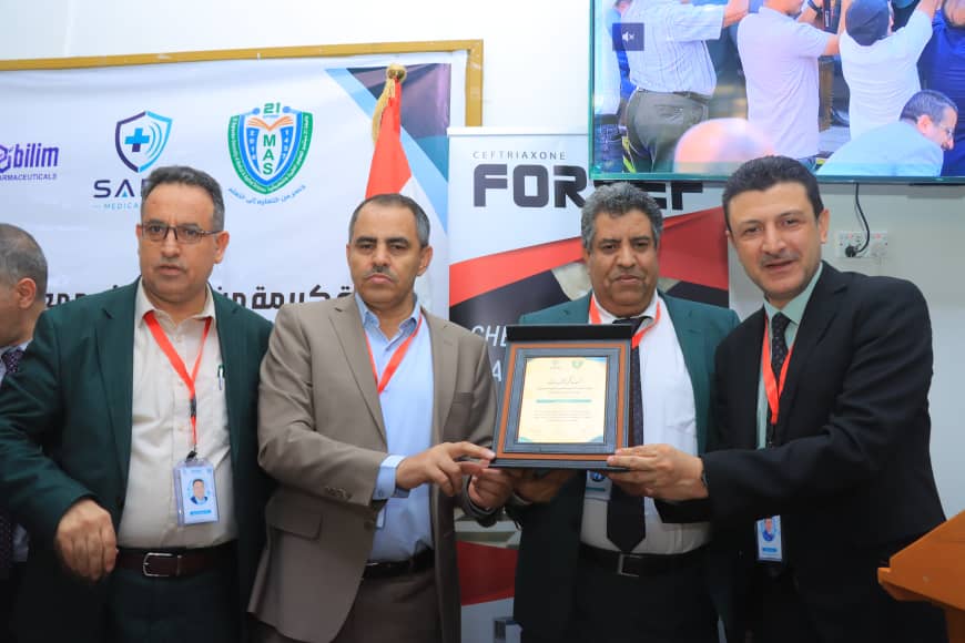 Group of four men in suits at an award ceremony, a framed certificate being handed over amid banners and a TV screen in the background.