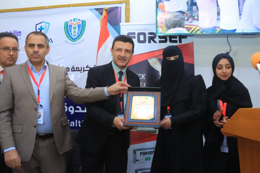Award ceremony: man in a suit presents a framed plaque to a woman in a black abaya, with another woman in hijab nearby and event banners in the background