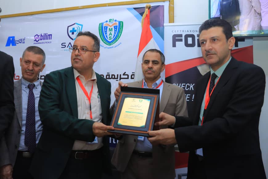 Men in suits at an awards ceremony exchange a framed certificate in front of event banners and a flag backdrop.
