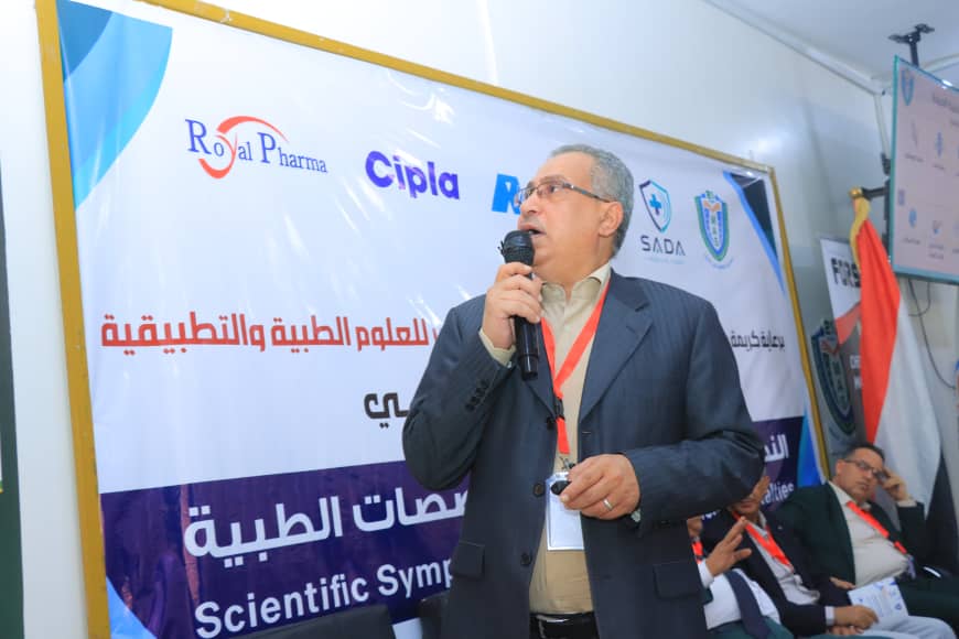 Man in a suit speaks into a handheld microphone at a medical/scientific conference, with banners and logos in the background.
