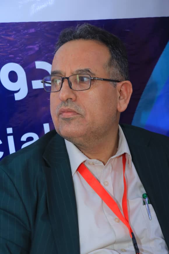 Middle-aged man wearing glasses and a dark pinstripe suit at a conference, with an orange lanyard behind him.