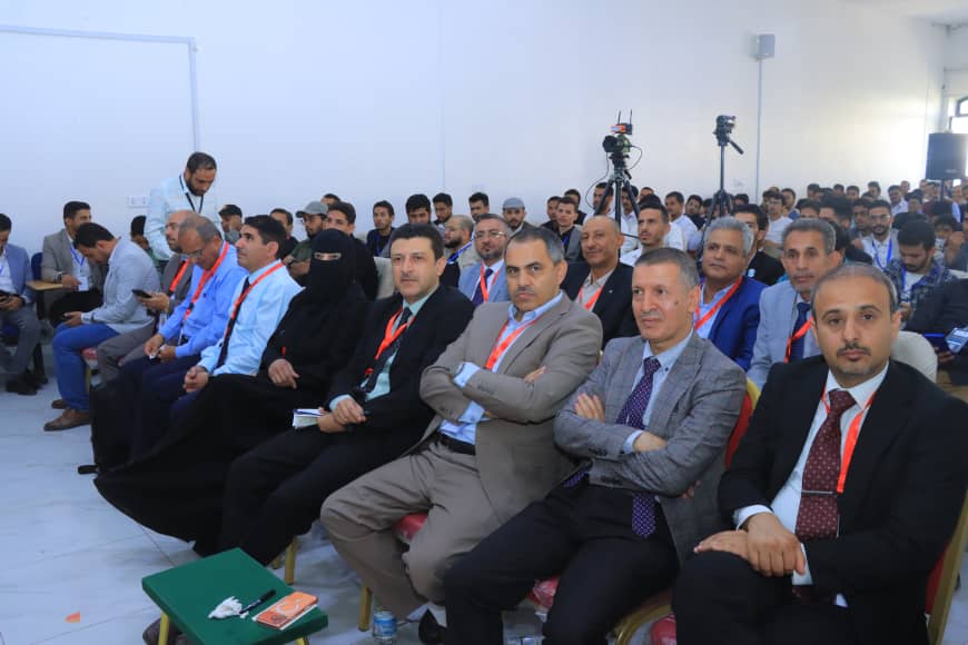 Crowded conference audience seated in rows, many men in suits with red lanyards.