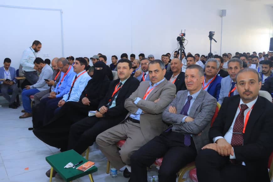 Audience seated in rows at a formal conference, many men in suits with red lanyards; two cameras on tripods in the back and a few attendees taking notes or looking at devices.