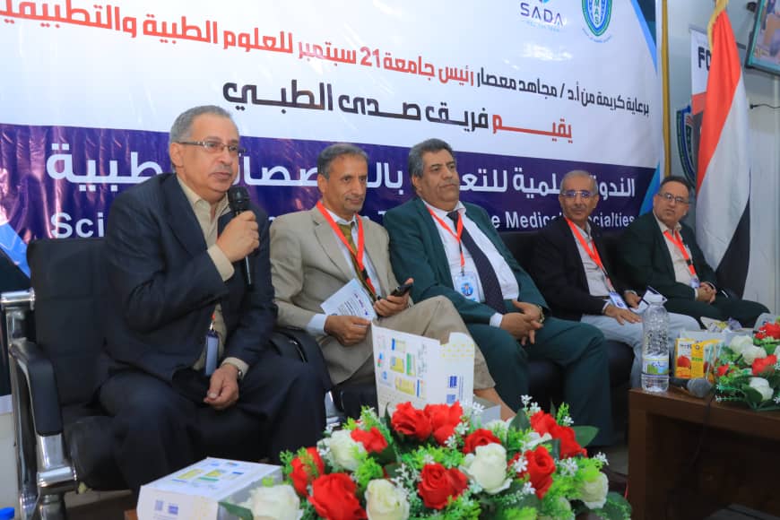 Five men in suits sit on a conference panel with Arabic banners behind them and flowers on the table.