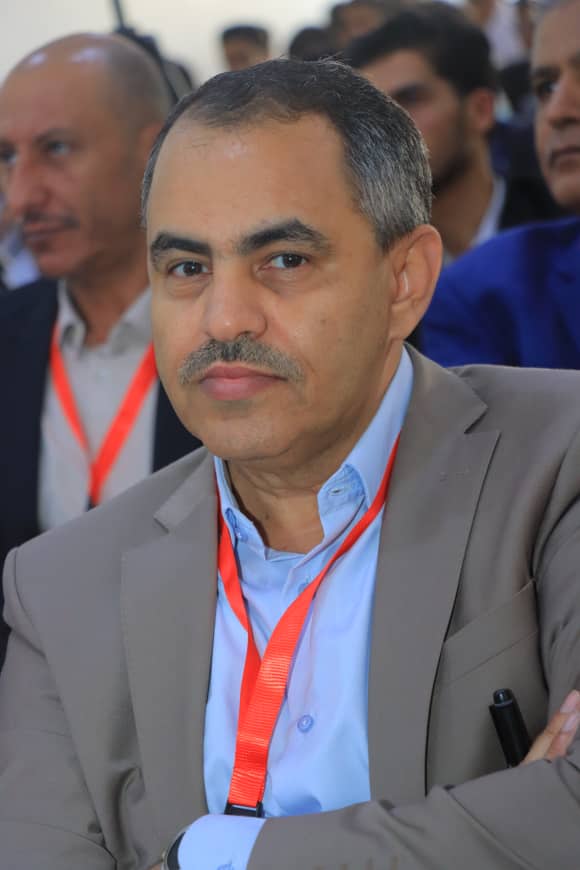 Portrait of a man in a gray suit and blue shirt wearing a red conference lanyard at an event.