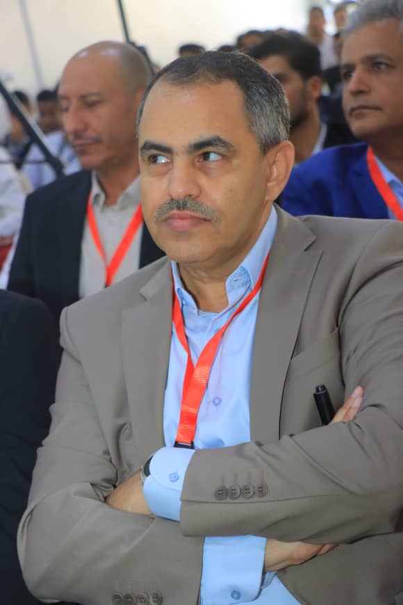 Middle-aged man in a beige suit with a blue shirt and orange lanyard sits with arms crossed at a conference, others in the background.
