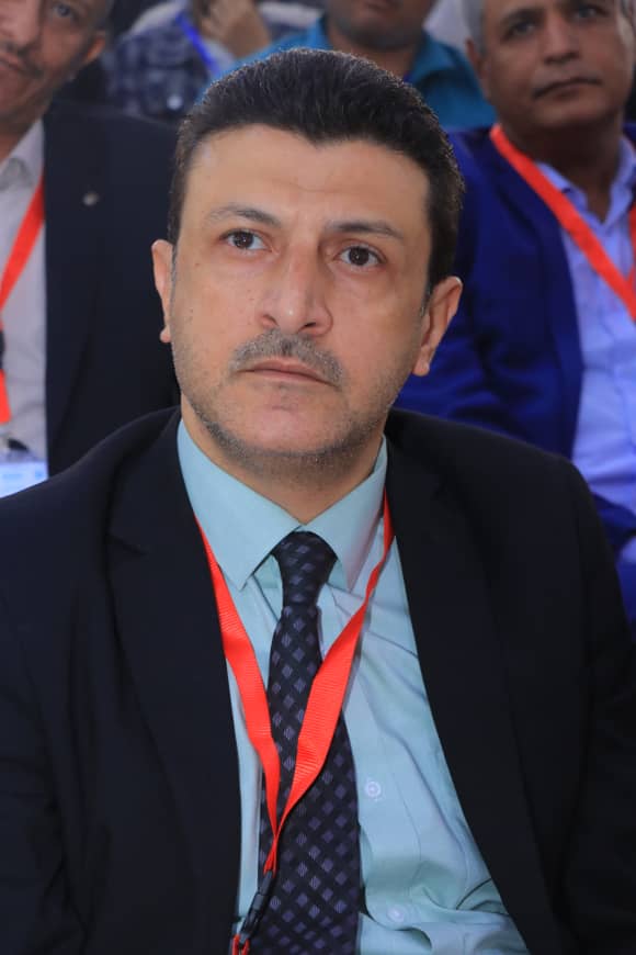Man in a dark suit and blue shirt wearing a red conference lanyard, looking ahead at a formal event.