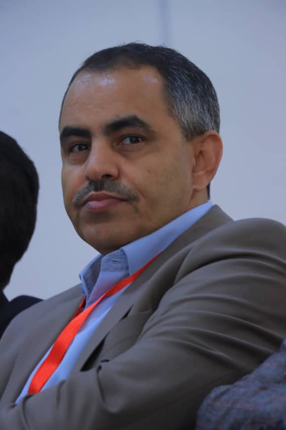 Middle-aged man in a gray suit and blue shirt wearing a red lanyard, seated at an event or panel discussion commonly used in conferences