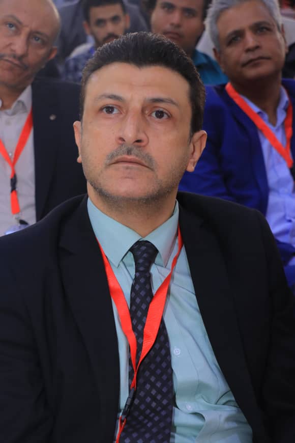 Conference attendee in a dark suit and light blue shirt with a red lanyard, looking slightly upward, seated among a crowd.
