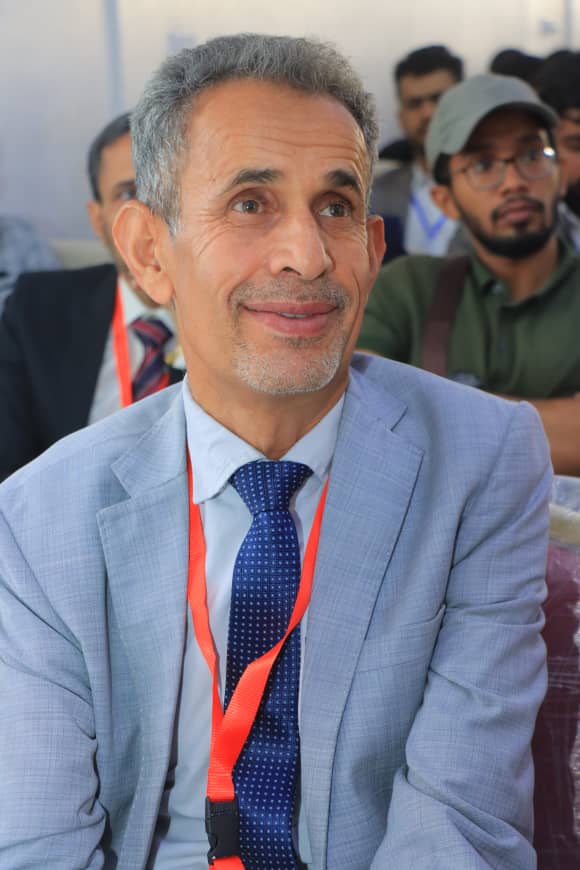 Man in a light blue suit and blue polka-dot tie wearing a red lanyard at a conference, smiling and looking to the side.