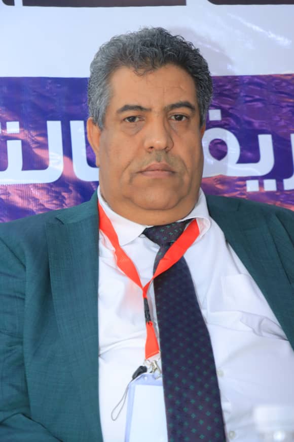Middle-aged man in a teal suit with a white shirt and navy tie, wearing a red lanyard and badge at a conference backdrop.