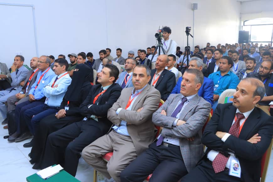 Conference audience seated in rows at a formal event, attendees wearing lanyards, with two camera operators filming in the back.