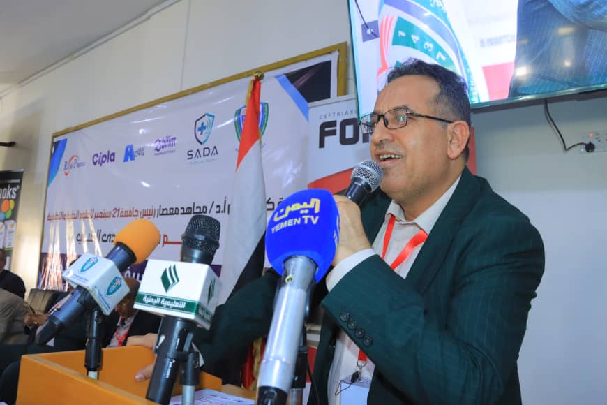 Man in a dark suit speaking into a microphone at a press conference, surrounded by several branded microphones and a banner backdrop with flags and logos.