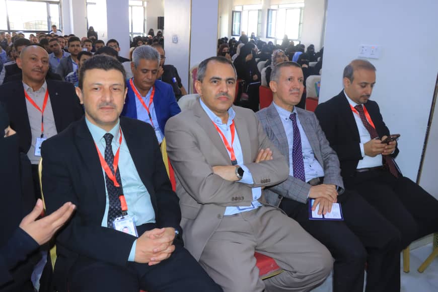 Group of men in suits with red lanyards seated in a conference hall, foreground attendees looking toward the stage.