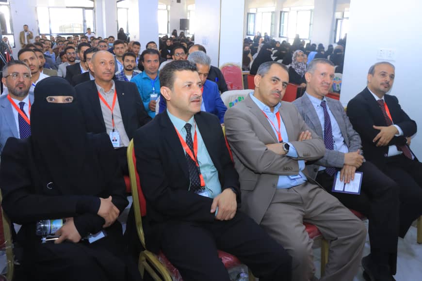 Audience seated in a bright conference hall, front row includes a woman in a black abaya and several men in suits wearing red lanyards.