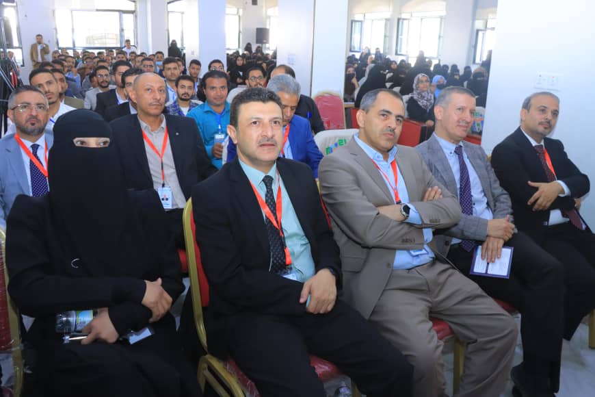 Front row of a conference audience: a woman in a black niqab sits among men in suits with red lanyards; a crowded hall behind them.