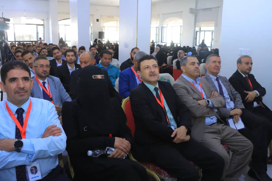 Crowded conference room with attendees seated and watching a presentation, including a woman wearing a black niqab in the front row.