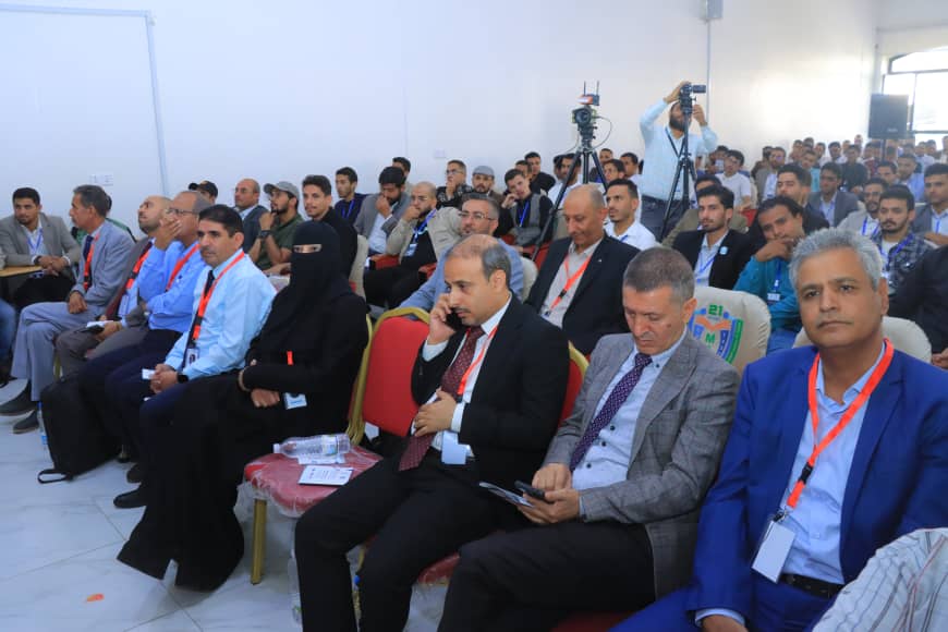 Large conference audience seated in a bright room, many wearing suits with lanyards; a man in a dark suit calls on his phone in the foreground.