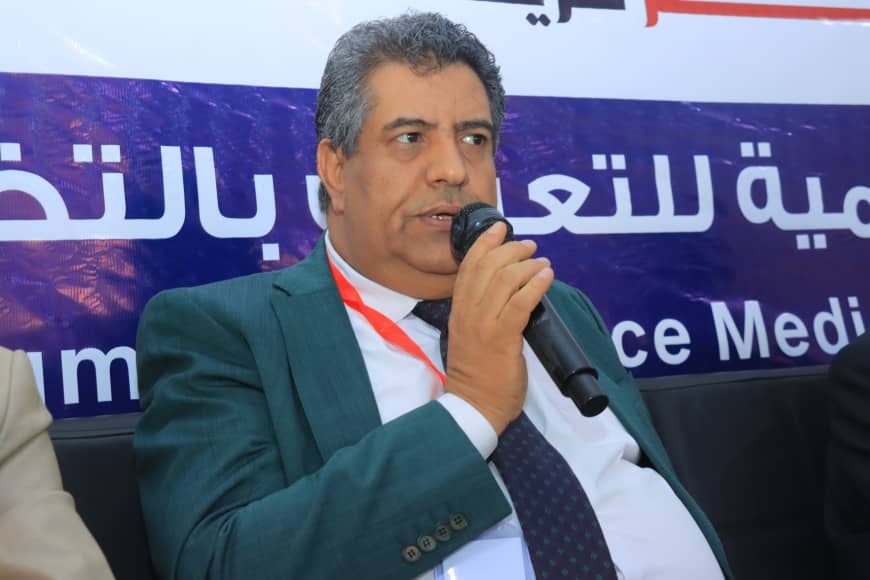 Man in a teal suit speaking into a microphone at a conference, with a blue banner in the background displaying Arabic text.