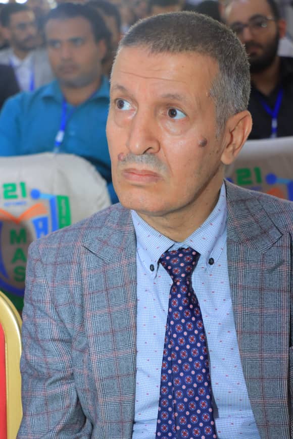 Older man with short gray hair in a gray plaid blazer and blue shirt, wearing a patterned tie, seated at a conference with a neutral expression and blurred audience in the background.