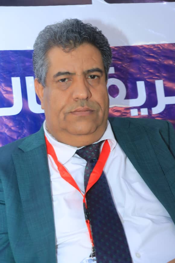 Man in a teal suit and white shirt wearing a red lanyard, sitting at an event with a purple banner in the background.