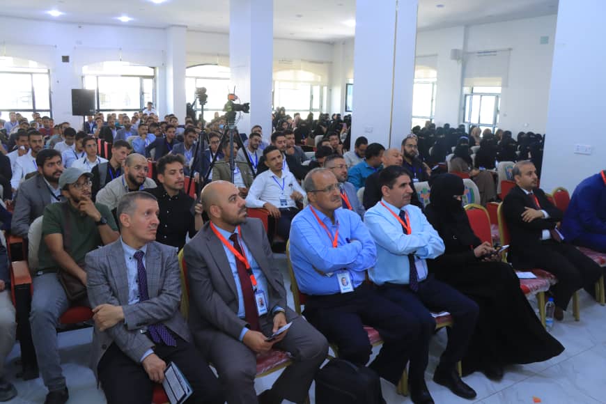Audience seated in a conference hall listening to a presentation, with attendees wearing badges and a camera on a tripod in the background.