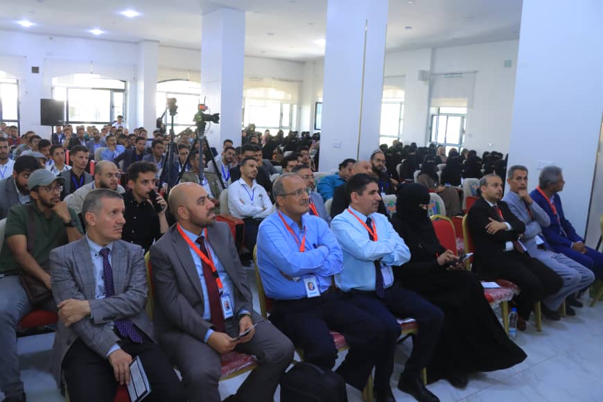 Audience seated in a bright conference room watching a presentation, front-row attendees in suits with lanyards and a woman in a black hijab visible on the right.