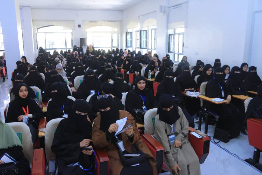 Large lecture hall filled with women wearing black abayas and niqabs, seated in rows for a conference or seminar, many with lanyards and note papers.