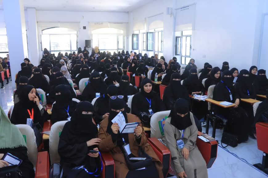 Large conference hall full of women in black hijabs seated in rows, many with blue lanyards, attending a seminar.