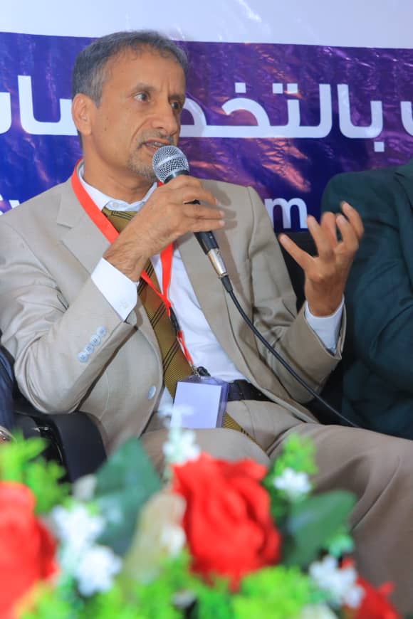 Man in a beige suit speaks into a handheld microphone at a panel, wearing a red lanyard and badge with a floral foreground.