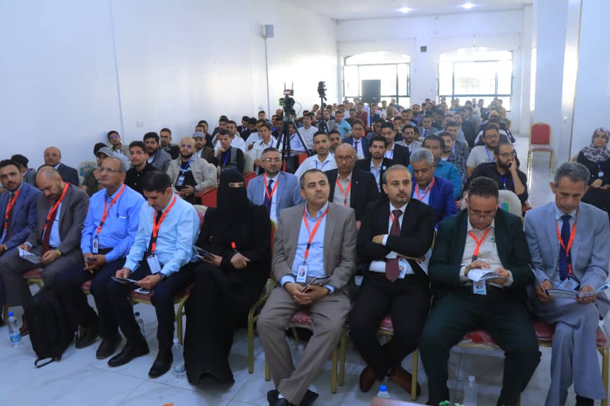 Large audience in a bright conference hall wearing red lanyards, seated for a presentation.