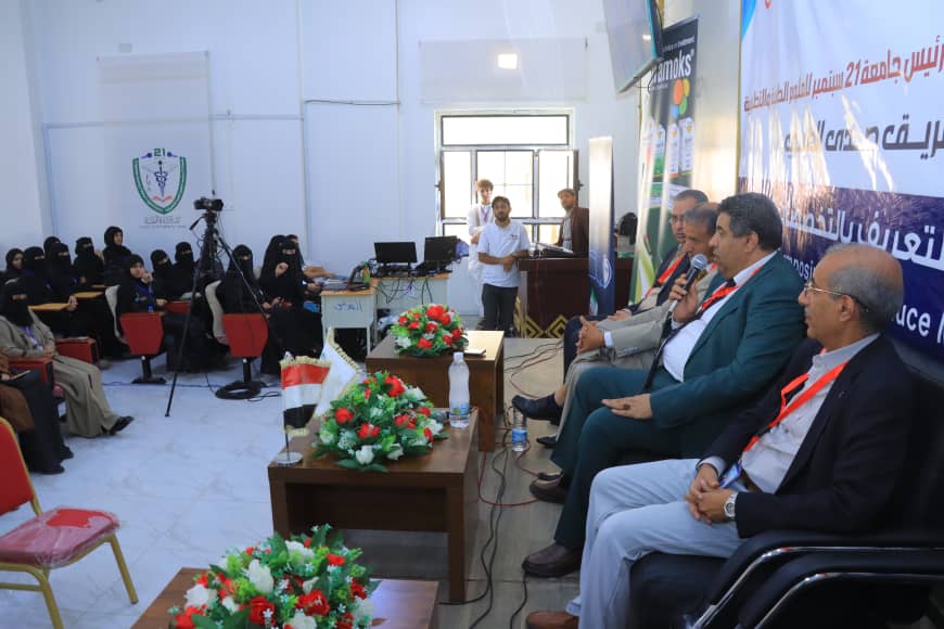 Panel discussion with four men seated on a stage, banners and flower arrangements behind them, audience seated in a hall with cameras and a banner in Arabic on the wall.