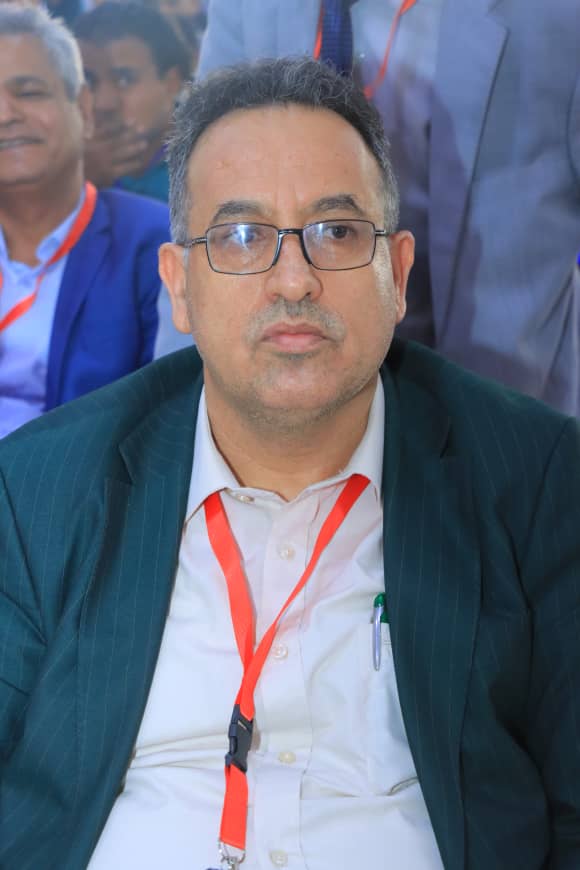 Man wearing glasses and a dark green pinstriped suit with a white shirt, red lanyard around his neck at a conference or event, seated in front of a backdrop with people behind him.