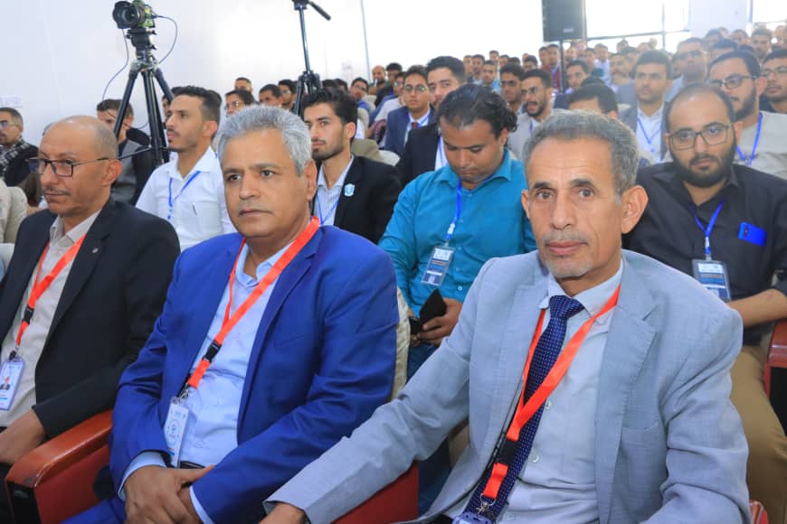 Audience at a conference: three men in the front row in suits with orange lanyards, seated in a lecture hall with a camera on a tripod and many attendees in the background.