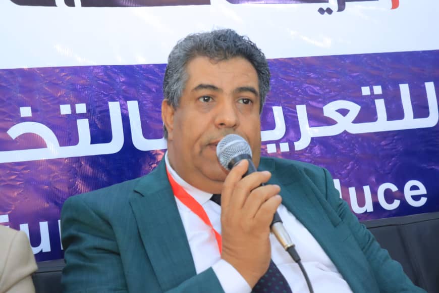 Middle-aged man in a teal suit speaks into a handheld microphone at a conference, with a purple banner in the background.
