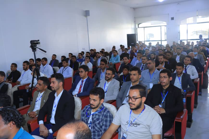 Large conference room filled with men wearing lanyards, seated for a presentation; a camera on a tripod records the event.