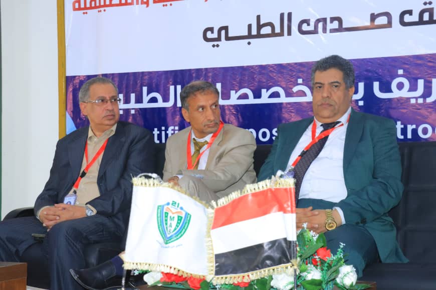 Three men in suits sit on a panel stage during a conference, with a purple Arabic banner behind them and Iraqi flag on a table in front.