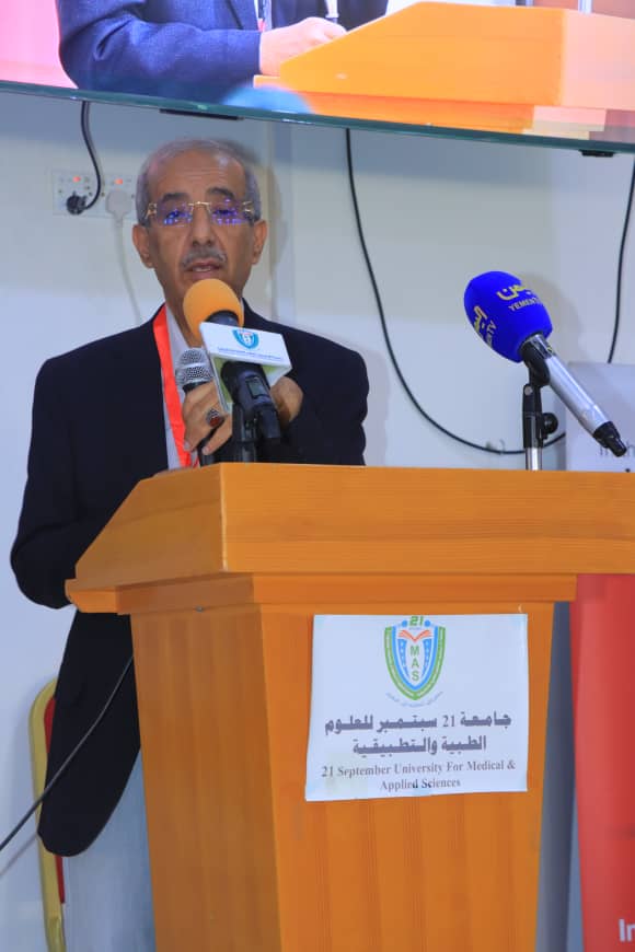 A man in a dark suit speaks at a wooden podium with two microphones during a university event; sign on the podium reads '21 September University for Medical & Applied Sciences'.