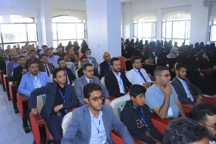 Audience seated in a conference hall; front rows in suits, many men with lanyards, and women in hijabs seated in the back.