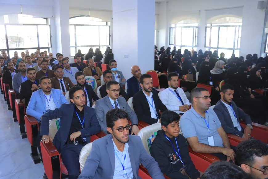 Audience seated in a bright conference hall, many wearing blue lanyards, with men in suits in the front rows and women in hijabs in the back.