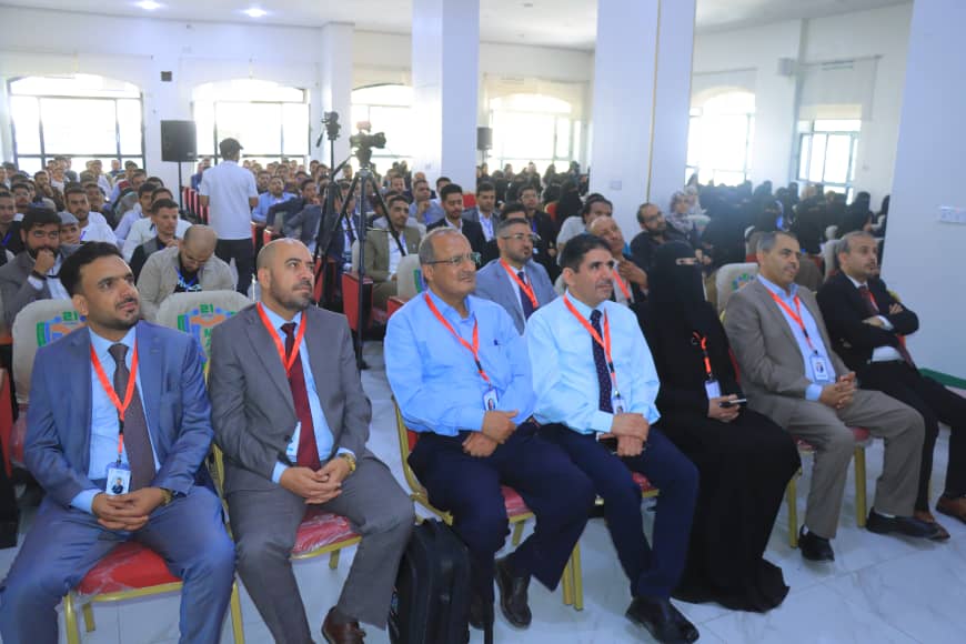 Audience seated in a conference hall, front rows wearing suits and red lanyards, with a camera on a tripod in the background.