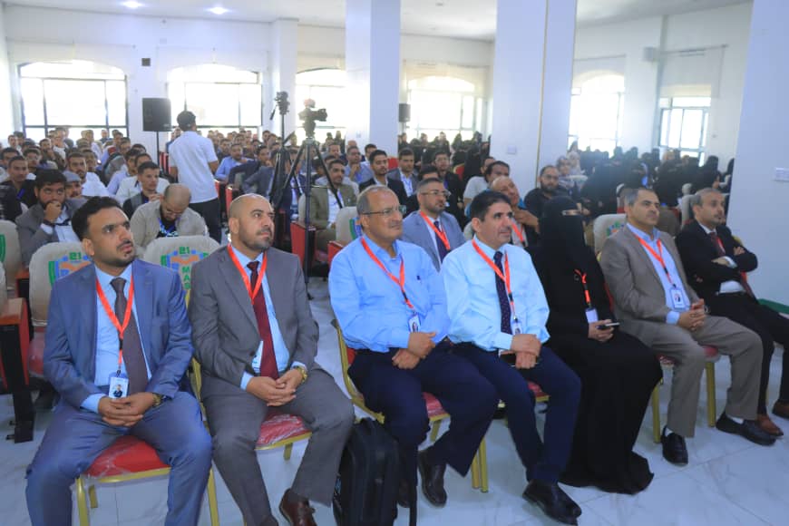 Audience seated in a bright conference hall, front-row men in suits wearing orange lanyards and ID badges, attentively listening to a presentation.